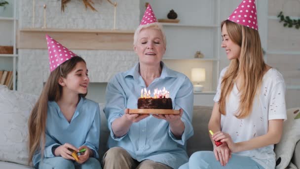 Happy vzrušený krásný tři ženy věk generace rodina stará babička mladá maminka a malé dítě vnučka slaví narozeniny ve party klobouky gratulujeme společně. Babička fouká svíčky - Záběry, video