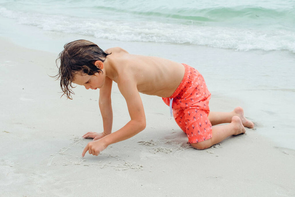 Παιδικό παιχνίδι σε τροπική παραλία. Το παιδί παίζει στη θάλασσα στις καλοκαιρινές οικογενειακές διακοπές. Τα παιδιά διασκεδάζουν στο παραθαλάσσιο θέρετρο. Αστείο αγόρι γράφει στην άμμο.Ενεργός τρόπος ζωής των παιδιών, κολύμπι οι άνθρωποι - Φωτογραφία, εικόνα