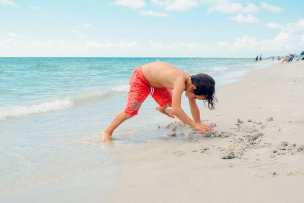 Lapsi leikkii trooppisella rannalla. Poika leikkii meressä kesäperhelomalla. Lapset pitävät hauskaa rantalomakohteessa. Hauska poika kirjoittaa hiekkaan.Aktiivinen lasten elämäntapa, uivat ihmiset - Valokuva, kuva