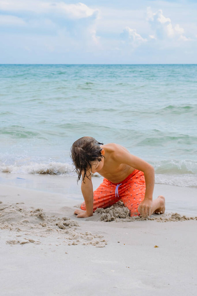 Παιδικό παιχνίδι σε τροπική παραλία. Το παιδί παίζει στη θάλασσα στις καλοκαιρινές οικογενειακές διακοπές. Τα παιδιά διασκεδάζουν στο παραθαλάσσιο θέρετρο. Αστείο αγόρι γράφει στην άμμο.Ενεργός τρόπος ζωής των παιδιών, κολύμπι οι άνθρωποι - Φωτογραφία, εικόνα