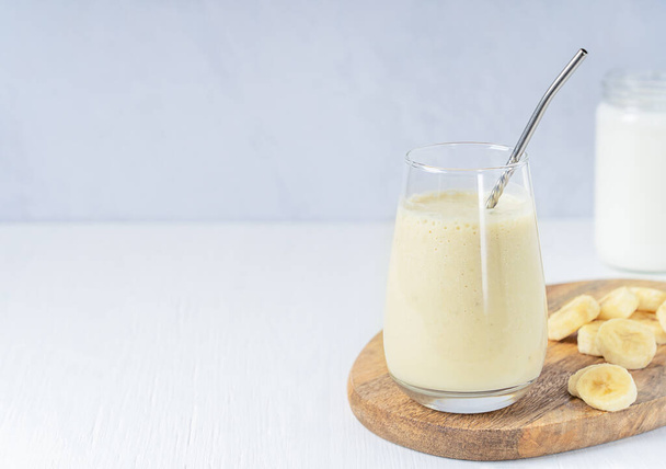 Indian lassi drink or milkshake made of ripe sweet bananas blended with milk served in drinking glass with metal straw and fruit slices on cutting board on white wooden table. Image with copy space - Foto, afbeelding