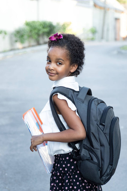 Happy school-age child with a backpack holding an orange folder. Portrait of cheerful mixed race girl. Afro hair. Back view. Outdoors. Back to school, childhood, schoolchildren, education concept - Foto, Imagen