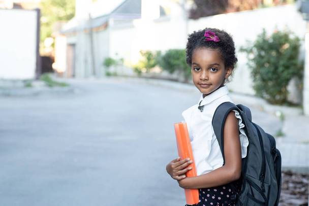 Heureux enfant d'âge scolaire avec un sac à dos tenant un dossier orange. Portrait de joyeuse fille métissée. Cheveux afro. En plein air. Retour à l'école, enfance, écoliers, concept éducatif Copier l'espace. - Photo, image