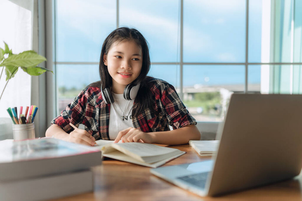 Asiatica teen girl indossa cuffie imparare la lingua online, utilizzando il computer portatile, guardando lo schermo, facendo compiti scolastici a casa, scrivere appunti, ascoltare lezioni o musica, istruzione a distanza - Foto, immagini