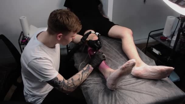Salón de tatuajes - el cliente acostado en el sofá y hacerse un tatuaje en la pierna en estilo graffiti - Metraje, vídeo