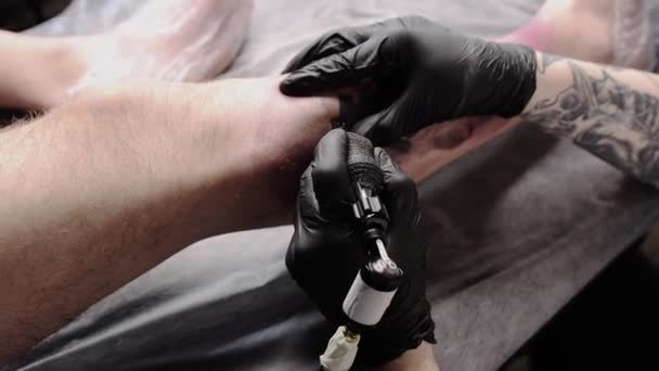 Salone del tatuaggio - il cliente sdraiato sul divano e con un tatuaggio sulla gamba in stile graffiti - il maestro riempie il tatuaggio con inchiostro nero - Filmati, video