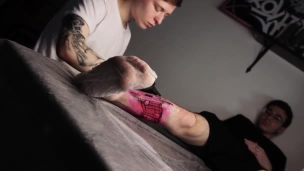 Salone del tatuaggio - cliente maschile sdraiato sul divano e con un tatuaggio sulla gamba - il padrone riempie il tatuaggio di inchiostro nero - Filmati, video