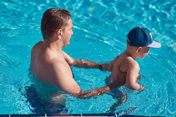 Padre e hijo caucásicos en la piscina. Padre le enseña a su hijo a nadar en la piscina en un día soleado. Pasando el rato juntos. Diversión de verano - Foto, Imagen