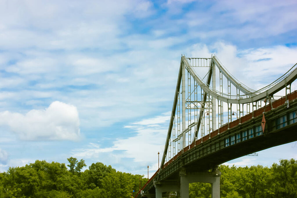 Yazın mavi bulutlu bir gökyüzüne karşı destekleyici yapısı ve feneri olan modern bir yaya demirinden yapılmış köprü. Ukrayna 'nın başkenti Kyiv' deki yüksek kırmızı bir köprüden görüntüyü al. Şehir mimarisi. - Fotoğraf, Görsel