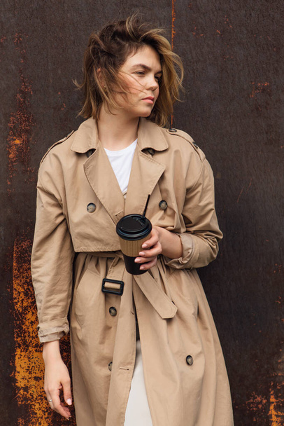 Young smiling millennial woman with wild hair dressed in an autumn coat standing a cup of coffee to go near the old rusty wall. - Foto, afbeelding