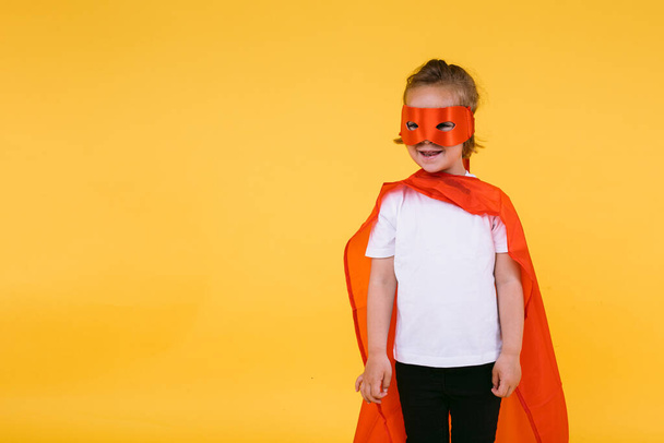 Petite fille blonde habillée en super-héroïne super-héros avec cape et masque rouge, souriante et regardant sur le côté, sur fond jaune - Photo, image