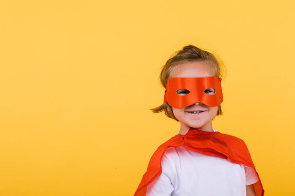Kleines blondes Mädchen im Superheldenkostüm mit Umhang und roter Maske, lächelnd, auf gelbem Hintergrund - Foto, Bild