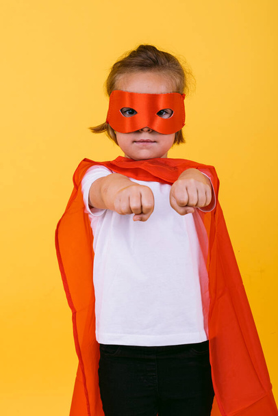 Little blonde girl dressed as a superheroine superhero with a red cape and mask, with arms in a flying position, on a yellow background - Photo, Image