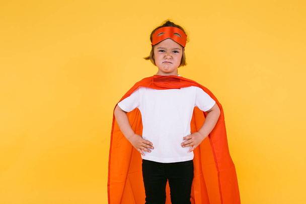 Bambina bionda vestita da supereroina supereroina con mantello e maschera rossa, arrabbiata, con le braccia akimbo serie, su sfondo giallo - Foto, immagini