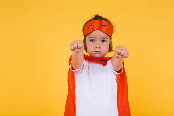 Petite fille blonde habillée en super-héroïne super-héros avec une cape rouge et un masque, les bras en position de vol, sur un fond jaune - Photo, image