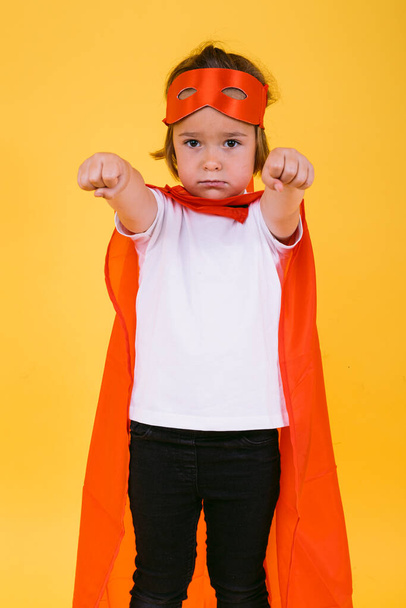 Little blonde girl dressed as a superheroine superhero with a red cape and mask, with arms in a flying position, on a yellow background - Photo, Image