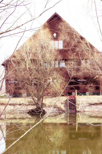 Photographie d'une belle maison entre des branches d'arbres nus. Maison de vacances à la campagne, chalet. Cabane en bois brun avec toit à pignon, escalier menant au bâtiment. Un étang devant la cour. Filtres chauds. - Photo, image