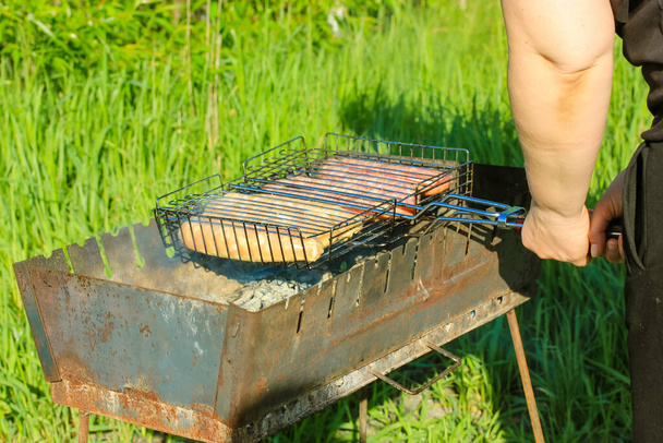 Raw sausage on a non-stick grill grid an the old metal BBQ. Preparing meat outdoors. Picnic in the woods on a sunny summer day. Green grass background. The process of cooking healthy food outdoors. - Photo, Image