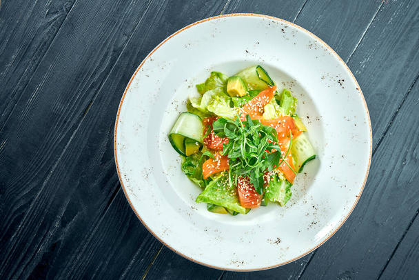 Appetizing, dietary salad with lettuce, cucumber, avocado and lightly salted salmon in a white plate on a dark wood background. Healthy food - Fotoğraf, Görsel