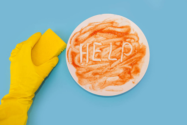 Top view photo of hands in yellow gloves holding a washcloth. White dirty plate with help sign on isolated blue background. Dishwashing concept. - Foto, Imagem