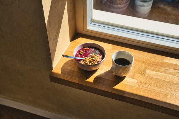 A high angle shot of a bowl of porridge with granola and almond nuts next to a mug of hot coffee on a windowsill - Φωτογραφία, εικόνα