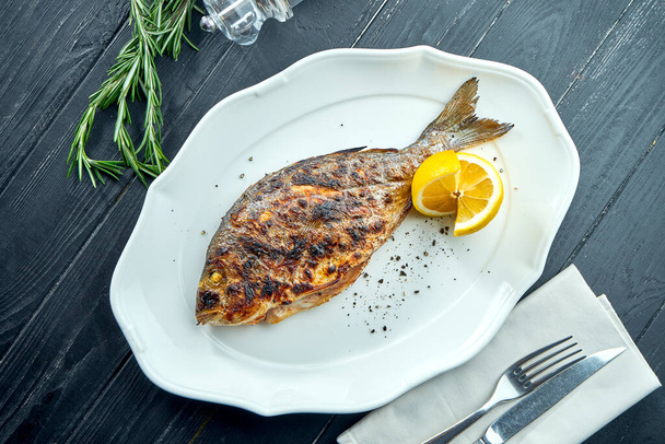 Appetizing grilled dorado on charcoal with lemon, served in a white plate on a dark wood background. Seafood BBQ - Foto, Imagen