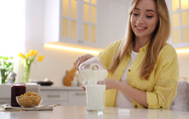 Young woman pouring milk from gallon bottle into glass at white table in kitchen - Photo, image