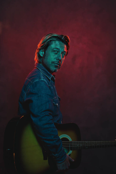 Man with acoustic western guitar in jeans shirt and jeans with belt in red and blue colored light. - Fotografie, Obrázek