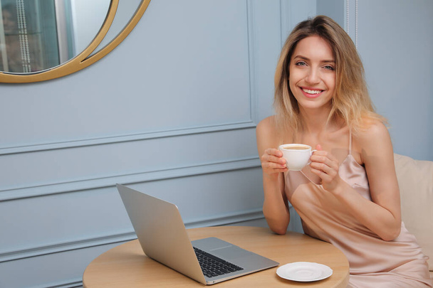 Happy young woman with cup of coffee and laptop at table indoors in morning - Foto, imagen