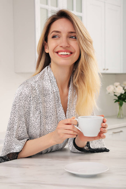 Pretty young woman in beautiful silk robe with cup of coffee at kitchen table - Foto, imagen