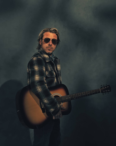 Man in checkered jacket and aviator sunglasses with acoustic western guitar in dappled sunlight in front of a grey wall. - Photo, Image