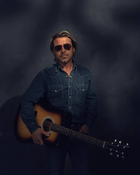 Man in denim shirt and aviator sunglasses with acoustic western guitar in dappled sunlight in front of a grey wall. - Photo, Image