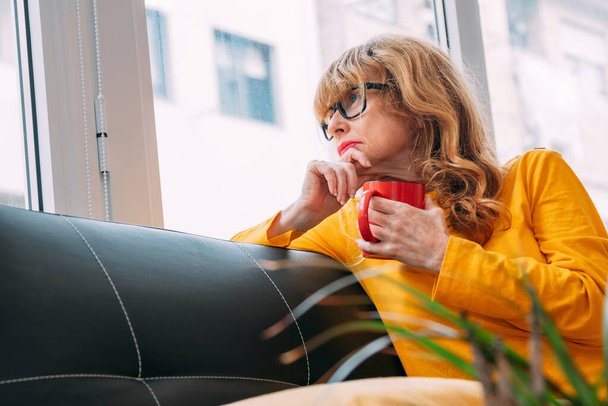 portrait of pensive or worried adult woman at home looking out the window with coffee cup - Photo, Image