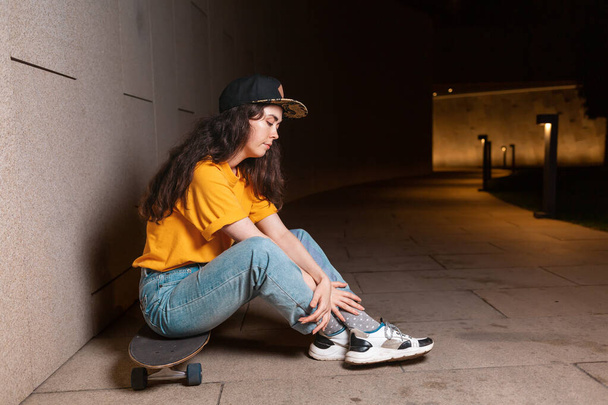 Una joven se sienta en un monopatín, posando en una calle nocturna. Al aire libre. Vista lateral. El concepto de subcultura juvenil. - Foto, Imagen