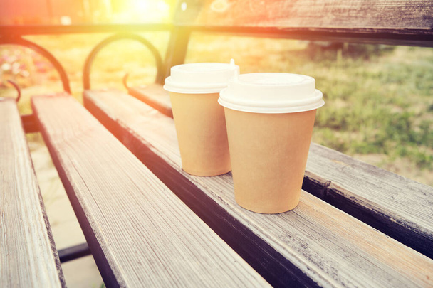 Two cups of coffee to take away in park on wooden bench in early morning with sunny glow - Фото, изображение