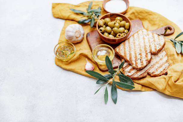 Green olives with olive oil with fresh baked ciabatta bread,  salt and young olive branches over white background. Flat lay, space - Foto, afbeelding