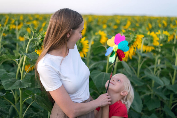 Young mom and son are walking in sunflower field with windmill toy. Green energy concept. Happy family. - Photo, Image