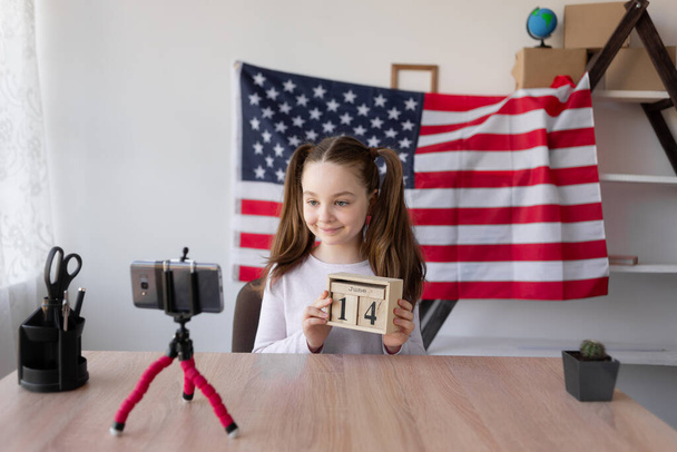 scolaretta si siede a casa a tavola sullo sfondo della bandiera americana. tiene un calendario in legno con la data del 14 giugno. Il giorno della bandiera americana. video blogger vicino a uno smartphone. - Foto, immagini