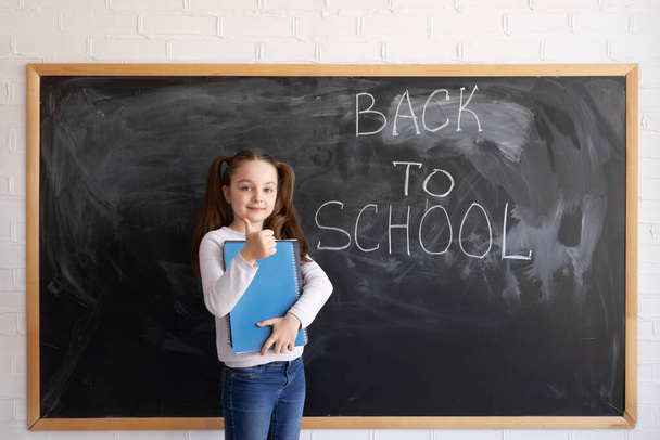 チョークボードを背景に若い白人の少女が立っている。その言葉は学校に戻って書かれている。手に大きなノートと親指を持ち. - 写真・画像