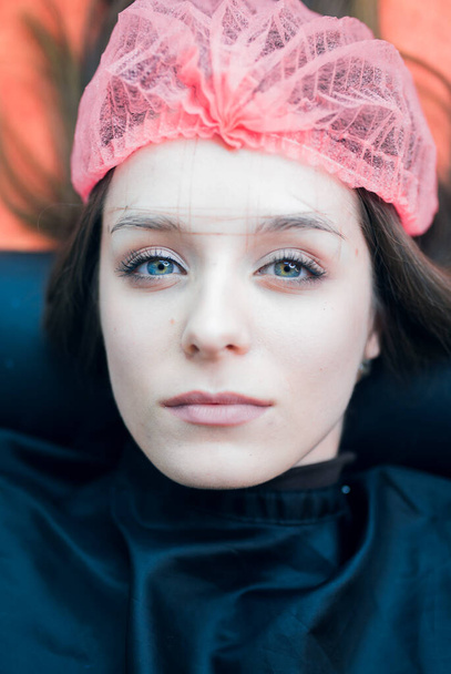 Das Porträt einer jungen Frau, die auf einem Bockbett liegt und eine Wischmütze mit Brauenumriss auf der Stirn und den Augenbrauen trägt. Permanente Make-up und Brauen machen professionelle Sphäre. - Foto, Bild