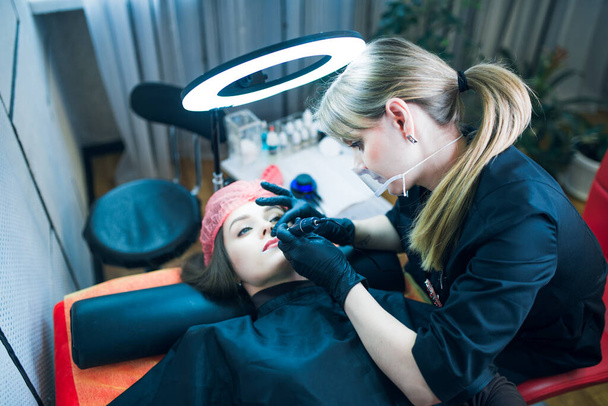 Maquillaje permanente artista en uniforme negro y guantes sostiene la máquina de tatuaje en los labios de su cliente con una lámpara borrosa, una silla y un equipo en el fondo. Cuidado de la piel, concepto de maquillaje permanente. - Foto, Imagen