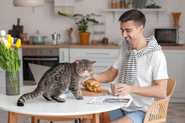 Jeune homme avec mignon chat manger croissant dans la cuisine - Photo, image