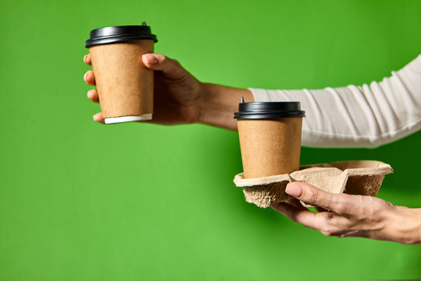 手は黒の蓋で2つのカップ茶色の紙を保持.2つのコーヒー特別オファーまたはプロモーション。緑色の背景に2つのカップを保持手。紅茶やコーヒーを移動します。黒蓋の茶色い紙コップ. - 写真・画像