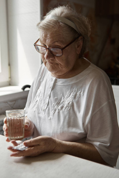 Грустная старуха, принимающая таблетки, проблемы со здоровьем в старости, дорогие лекарства. Руки пожилой женщины распаковывают несколько таблеток для приема лекарств. Бабушка принимает таблетки и пьет стакан воды - Фото, изображение