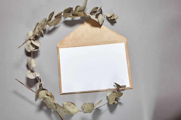 White paper sheet mockup on a brown craft envelope on gray background. White paper empty blank, branch of eucalyptus on gray table. Invitation card mockup. Flat lay, top view, copy space, mockup - Photo, Image