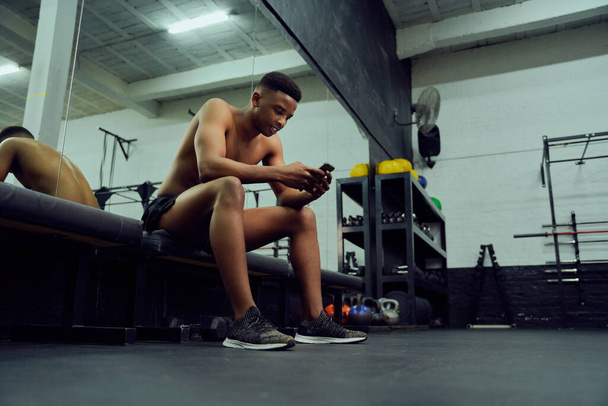 Young African American male looking at his phone to reply to an online message. Mixed race, male personal trainer looking at his phone while sitting down in the gym. High quality photo  - Foto, Imagen