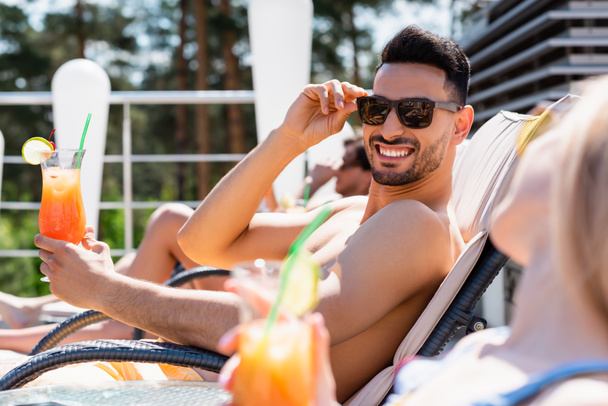 Homem árabe alegre em óculos de sol segurando coquetel perto de amigo borrado na cadeira deck  - Foto, Imagem