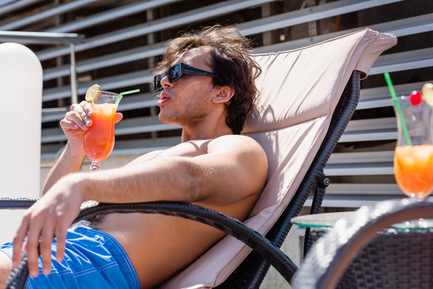 Homme aux lunettes de soleil tenant un cocktail tout en se relaxant sur une chaise longue au resort  - Photo, image