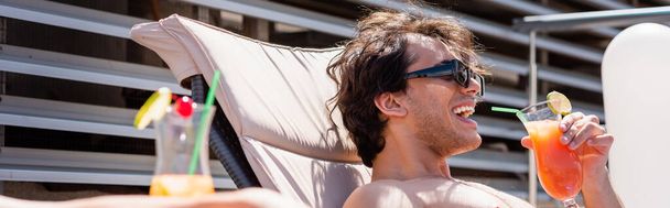 Hombre positivo en gafas de sol celebración de cóctel con paja para beber en la silla de cubierta, bandera  - Foto, Imagen