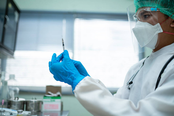 Mladý lékař v bílé uniformě s obličejovým a obličejovým štítem připraví injekční stříkačku pro očkování osob v lékařském středisku. - Fotografie, Obrázek